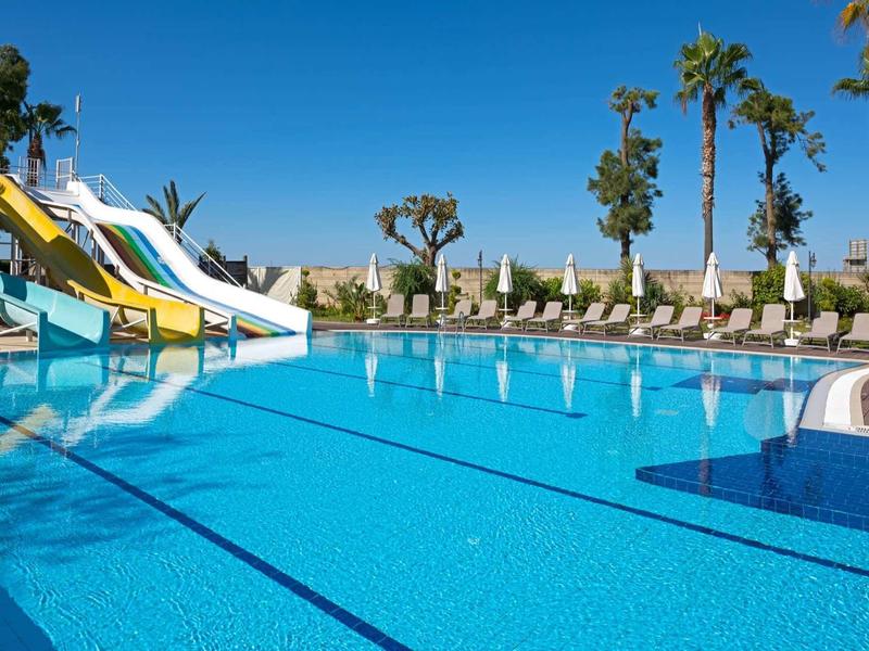 Piscina blu con scivoli, palme e ombrelloni sotto un cielo sereno.