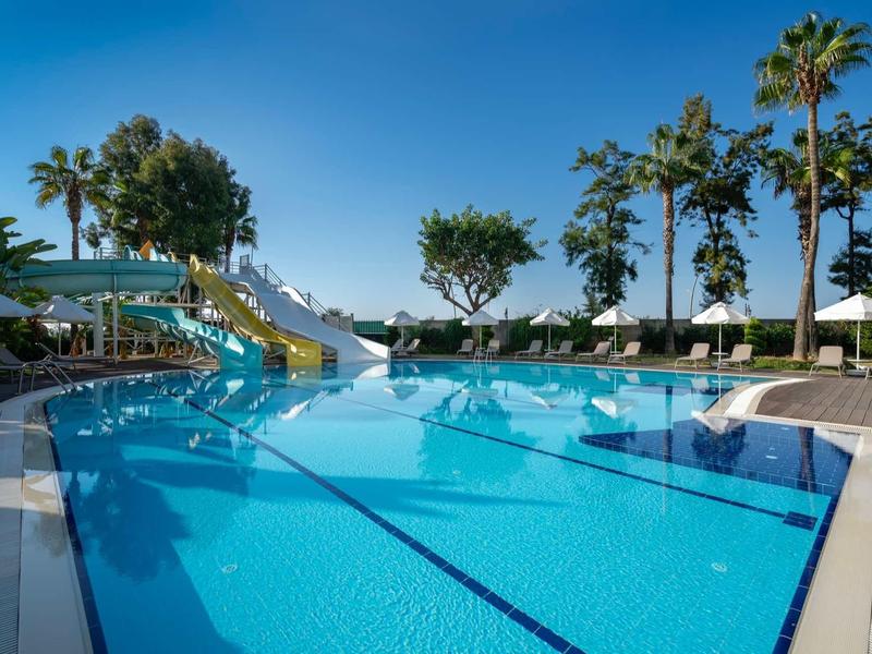Piscina all'aperto con scivolo, ombrelloni bianchi e palme sotto il cielo sereno.