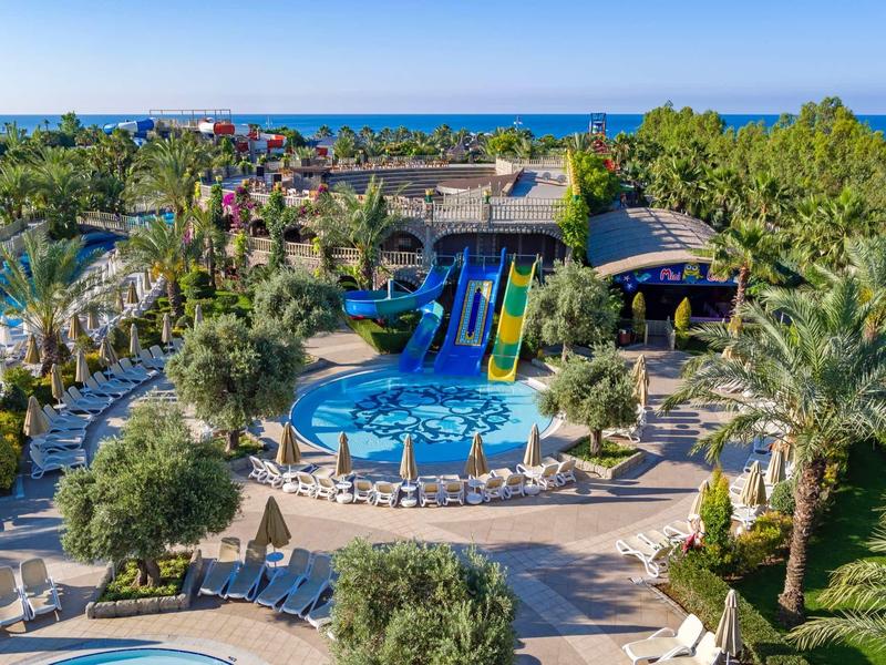 Complesso alberghiero con piscina, scivoli, palme e lettini sotto un cielo azzurro chiaro.