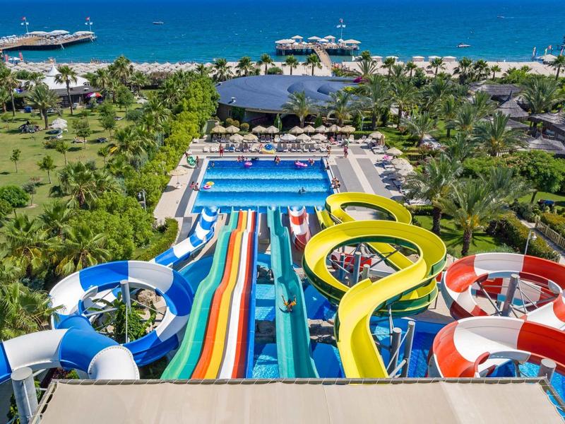 Ampio parco acquatico con scivoli colorati e piscina, palme e spiaggia sullo sfondo.