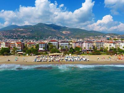 Strand mit türkisfarbenem Meer, Sand, Sonnenschirmen und dahinter Stadt und bewaldete Hügel.
