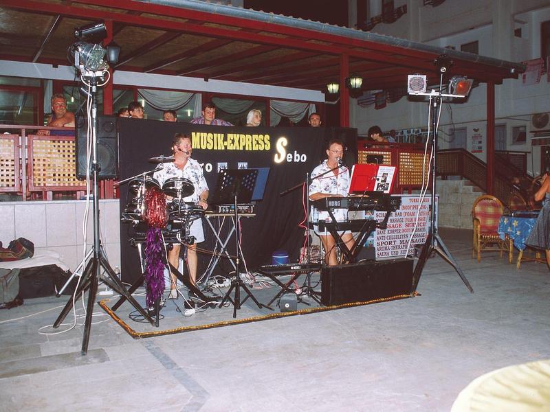 Piccola band che suona dal vivo al chiuso, circondata da luci e strumenti musicali.