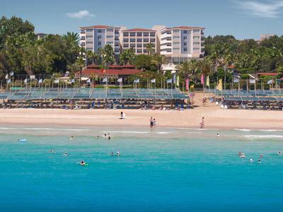 Blick auf einen Strand mit klarem, türkisfarbenem Wasser und einem großen Hotel im Hintergrund.
