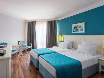 Helles Doppelzimmer mit zwei Betten, türkisfarbenen Akzenten, Schreibtisch und großem Fenster mit Vorhängen.