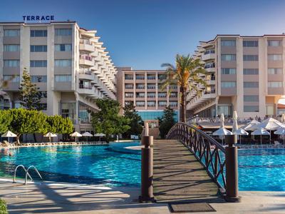 Hotel entrance with pool, wooden bridge, and umbrellas under clear sky.