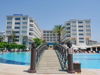 Hölzerne Brücke führt über einen Pool zu modernen weißen Hotelgebäuden unter blauem Himmel.