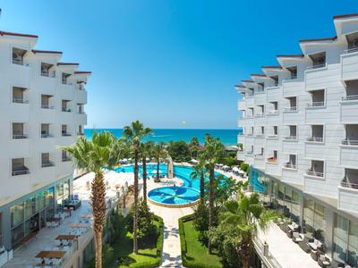 Hotel mit zwei weißen Gebäuden, Pool, Palmen und Blick auf das blaue Meer bei klarem Himmel.
