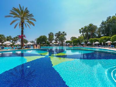 Großer Pool mit klarem blauem Wasser umgeben von Palmen und Sonnenschirmen unter blauem Himmel.