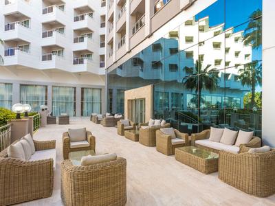 Moderne Außenlounge mit Korbmöbeln vor einem Hotel mit Glasspiegelung und Palmen.