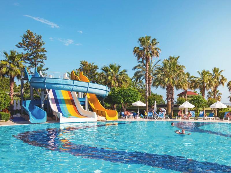 Parco acquatico con scivoli colorati, piscina e palme sotto un cielo sereno.