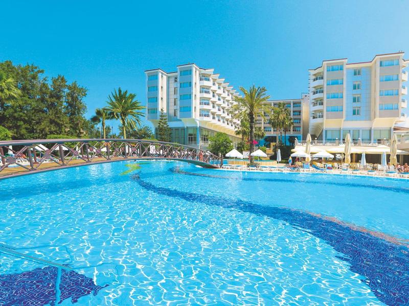 Grande piscina all'aperto davanti a un hotel moderno sotto un cielo azzurro limpido con palme.