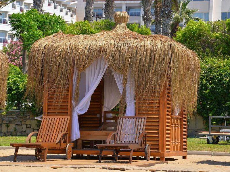 Capanna da spiaggia con tetto di paglia, tende e lettini in legno davanti a cespugli verdi e edifici