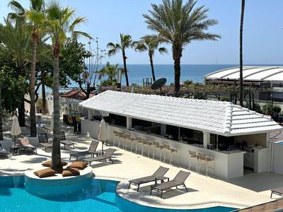 Moderner Poolbereich mit Liegen, Palmen und weißer Bar vor blauem Meer und klarem Himmel.