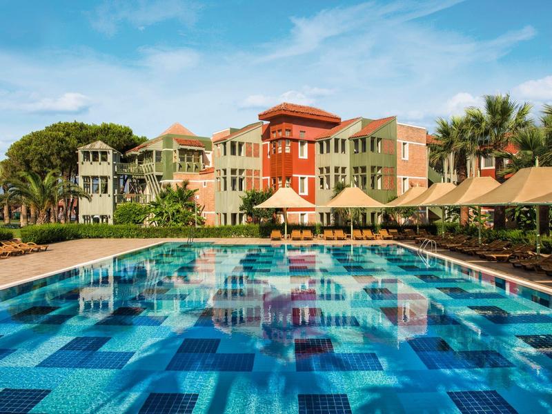Large hotel with blue tiled pool and palm trees under blue sky.