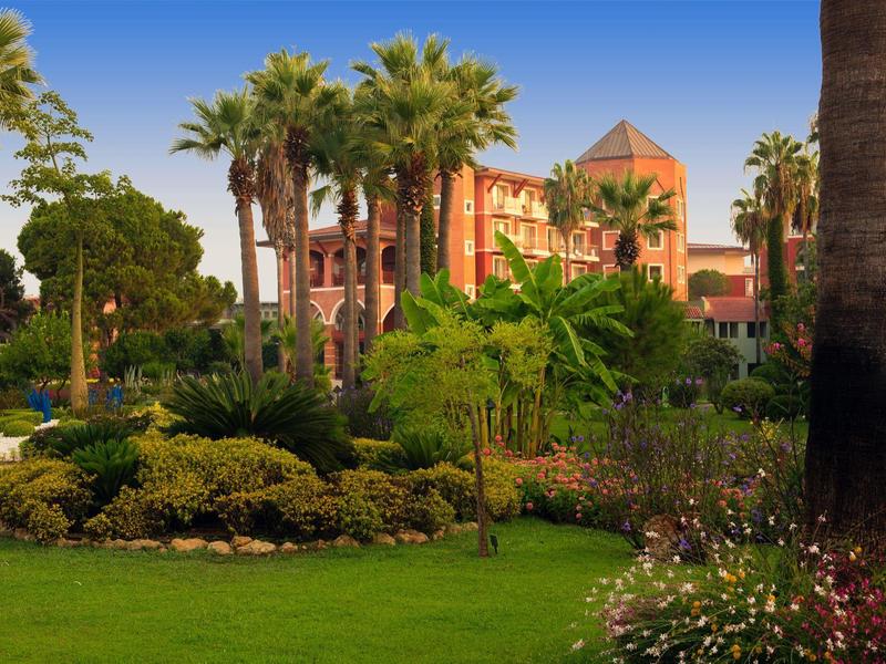 Lush garden with palm trees and bushes in front of a large hotel building at sunset.