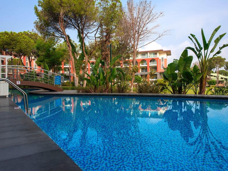 Clear blue pool surrounded by tropical plants and a hotel building in the background.