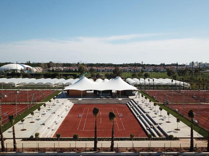 Outdoor tennis courts with a white canopy tent in the center and buildings in the background under a clear sky.