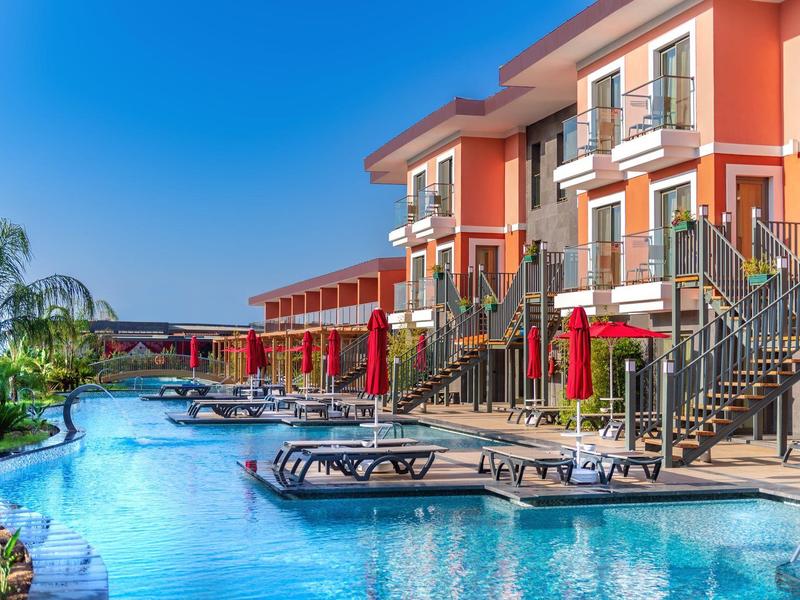 Luxury hotel pool area with sun loungers, stairs, balconies, and palm trees under blue sky.