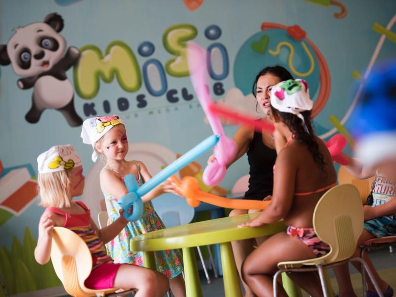 Children wearing party hats play with balloon swords at a small round table in a colorful playroom.