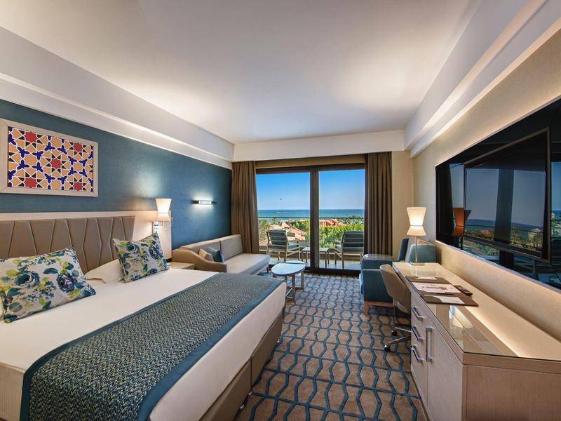 Modern hotel room with king bed, balcony overlooking pool and sea, and large TV.