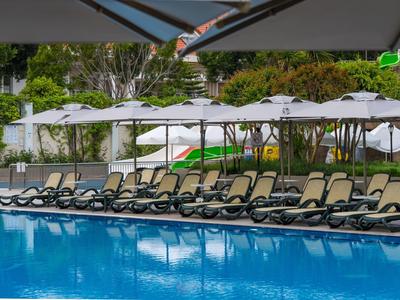 Mehrere Liegestühle und Sonnenschirme entlang eines blauen Pools in einer grünen Umgebung.