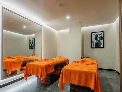 Modern spa room with two treatment beds, orange blankets, and a large wall mirror.