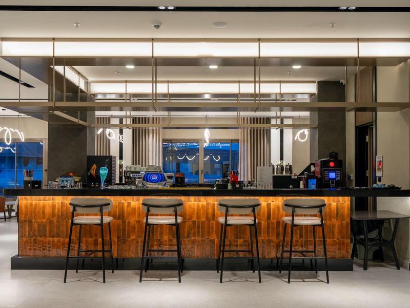 Modern hotel bar with wooden counter and black bar stools under warm lighting.