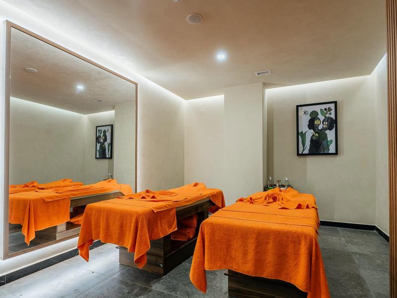 Modern spa room with two treatment beds, orange blankets, and a large wall mirror.