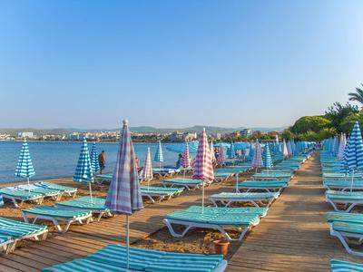 Strand mit blauen Liegen und bunten Sonnenschirmen entlang eines Stegs am ruhigen Wasser.
