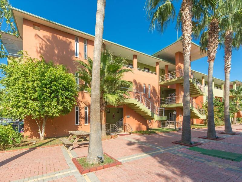 Sonnige Außenansicht eines zweistöckigen Hotels mit Palmen und einer gepflasterten Terrasse.