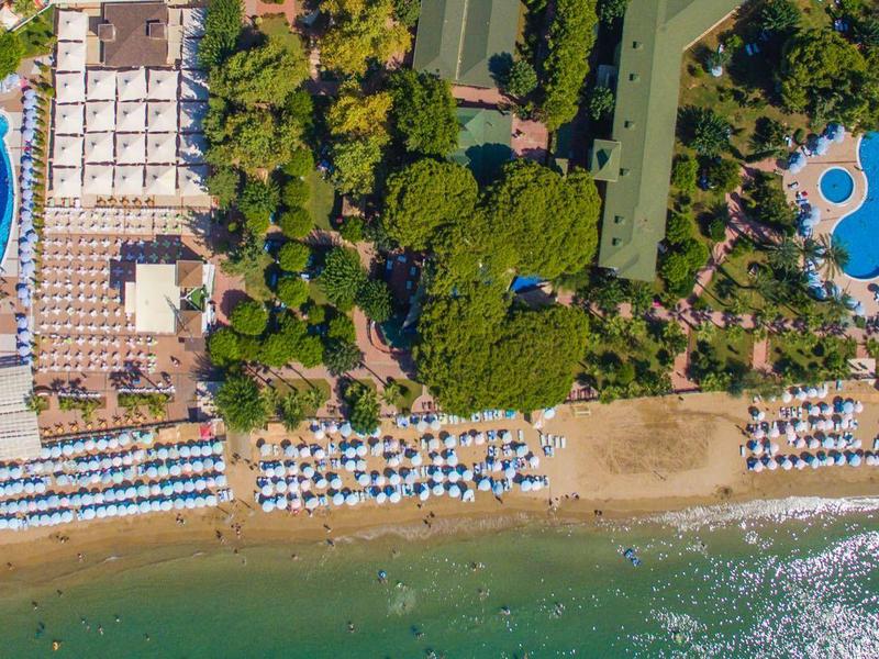 Luftaufnahme eines Strandes mit Liegen, Pools und viel grünem Bewuchs in einem Urlaubsresort.