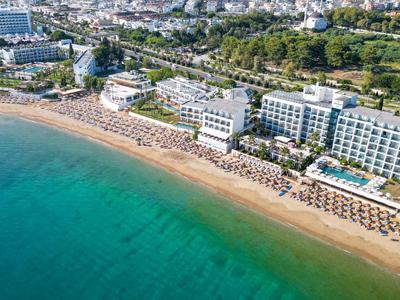 Vista aérea de un hotel junto a una playa de arena con sombrillas y mar azul.
