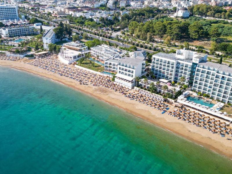 Vista aérea de un hotel junto a una playa de arena con sombrillas y mar azul.