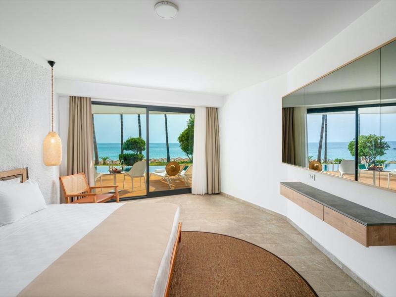 Habitación de hotel luminosa con gran ventana, vista al mar y terraza con muebles de jardín.