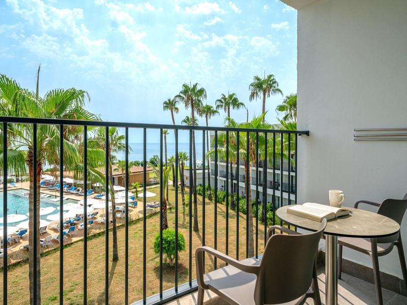 Balcón con mesa y sillas, vista a la piscina del hotel y palmeras bajo cielo azul.