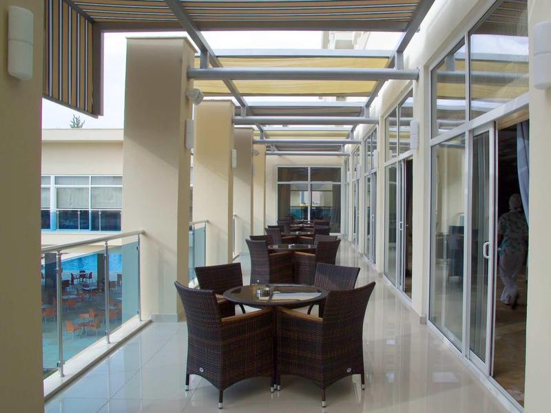 Moderner Balkon mit Glasgeländer, braunen Korbmöbeln und weißem Bodenbelag unter transparentem Dach.