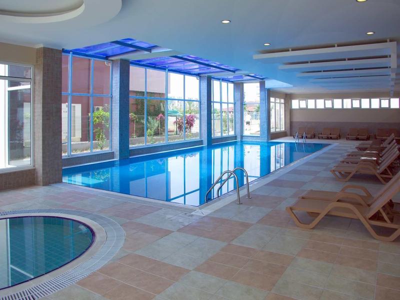 Innenraum mit großem Schwimmbecken, rundem Whirlpool, Liegestühlen und großen Fenstern.