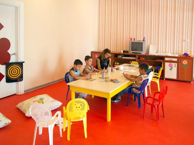 Kinder sitzen an einem gelben Tisch mit bunten Stühlen in einem Raum mit rotem Boden und heller Wand.