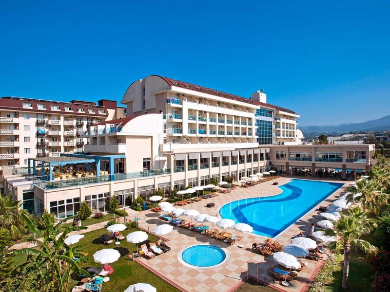 Großes Hotel mit blauem Pool, weißen Sonnenschirmen und Liegestühlen unter klarem Himmel.