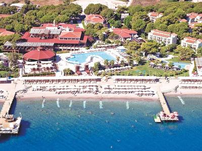 Luftaufnahme eines Strandes mit Liegen, zwei Stegen und dahinter Hotels in grüner Umgebung.