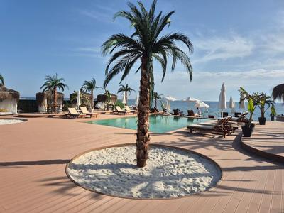 Poolbereich mit Palmen, Sonnenliegen und Sonnenschirmen unter blauem Himmel am Meer.