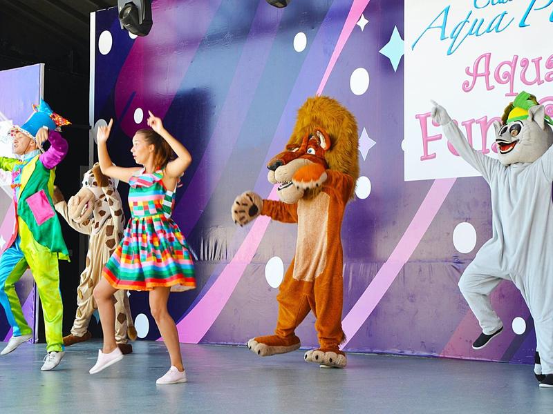 Děti a dospělí v barevných kostýmech tančí na pódiu s letní dekorací.