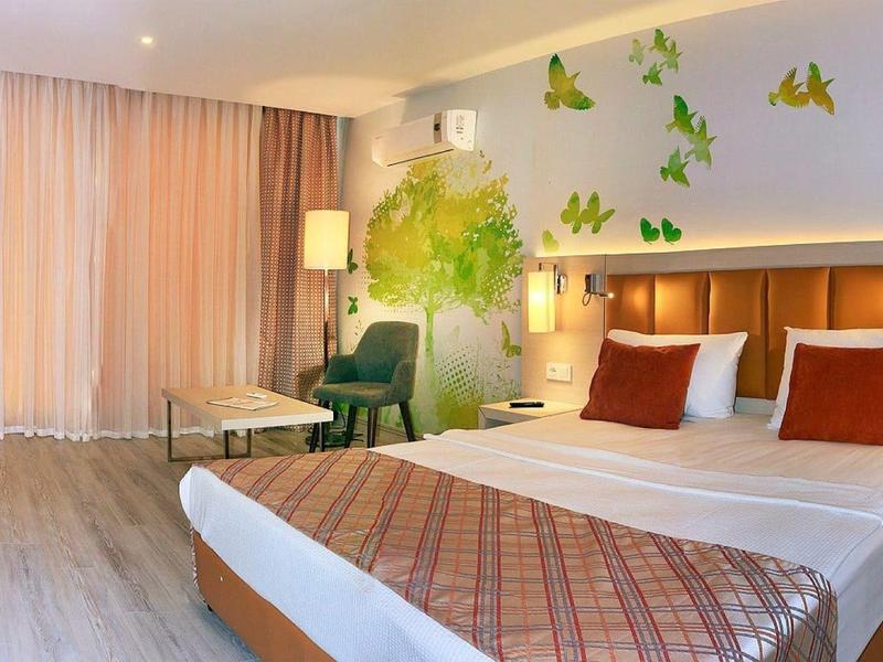 Moderní hotelový pokoj s manželskou postelí, zelenými křesly a motýlími nástěnnými dekoracemi.