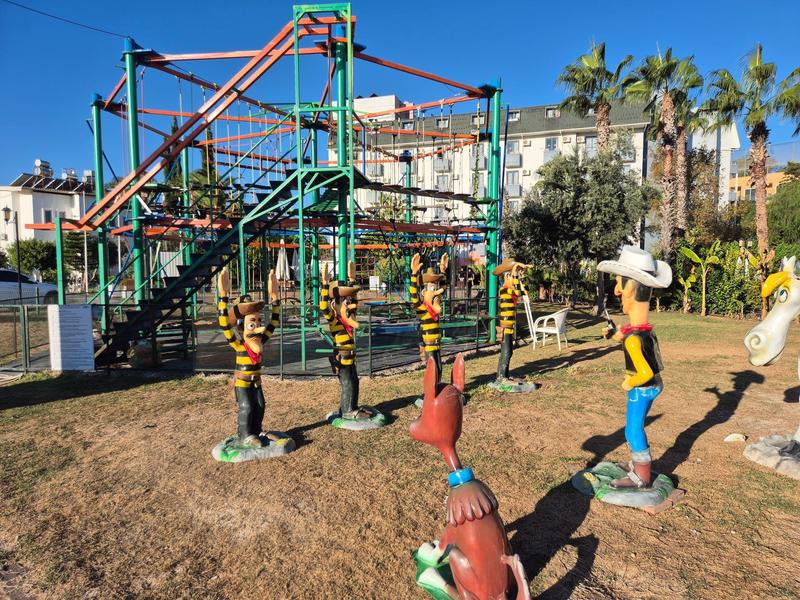 Děti si hrají na barevném lanovém skluzavce na slunném hřišti s palmami a budovami v pozadí