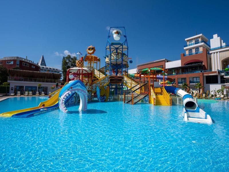 Vodní park s barevnými skluzavkami a čistým modrým bazénem před hotely pod jasnou oblohou.