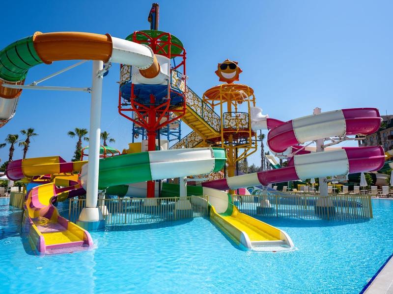 Barevné vodní skluzavky v venkovním bazénu za slunečné oblohy s palmami v pozadí.