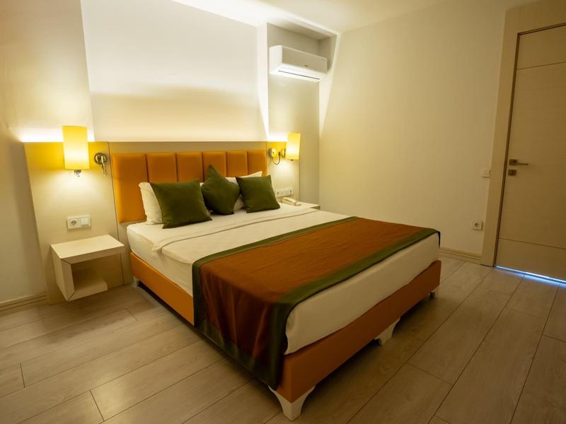 Příjemný hotelový pokoj s manželskou postelí, zelenými polštáři, teplým osvětlením a dřevěnou podlahou.