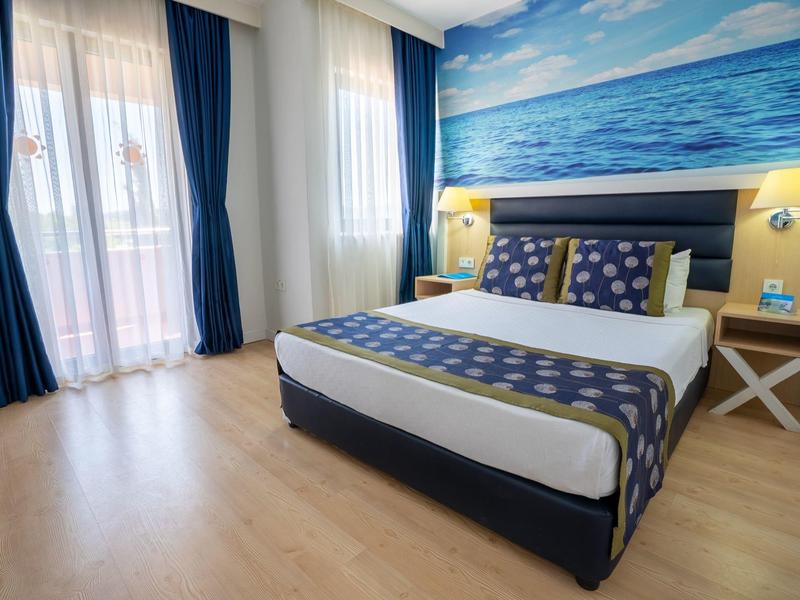 Světly hotelový pokoj s manželskou postelí, modrým ložním dekorem a velkými okny s záclonami.