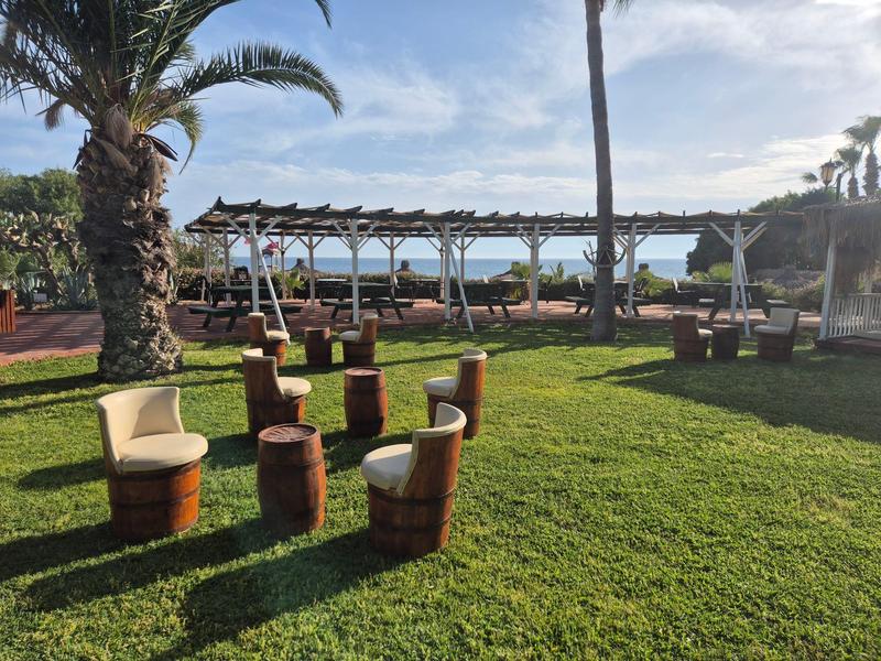 Zelená zahradní plocha s hnědými dřevěnými židlemi a bílými polštáři, palmy a výhled na moře