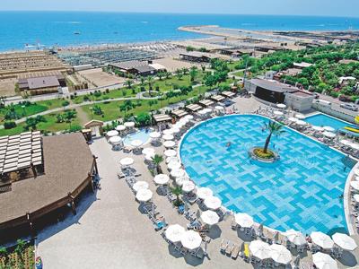 Großer blauer Pool mit Sonnenschirmen, mehreren Gebäuden und grünem Bereich am Meer im Hintergrund.